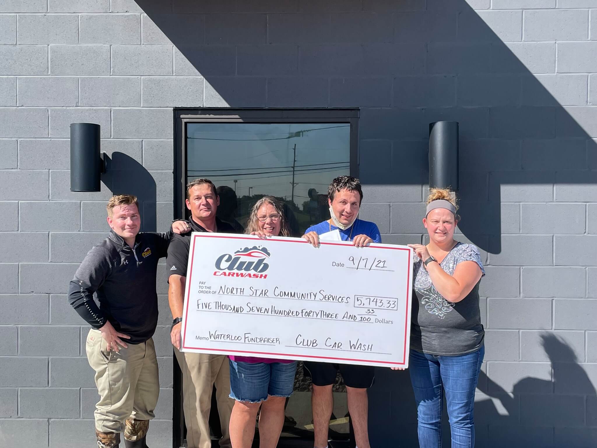 Club Car Wash employees and North Star Community Service representatives holding large check in front of the doors of the location.